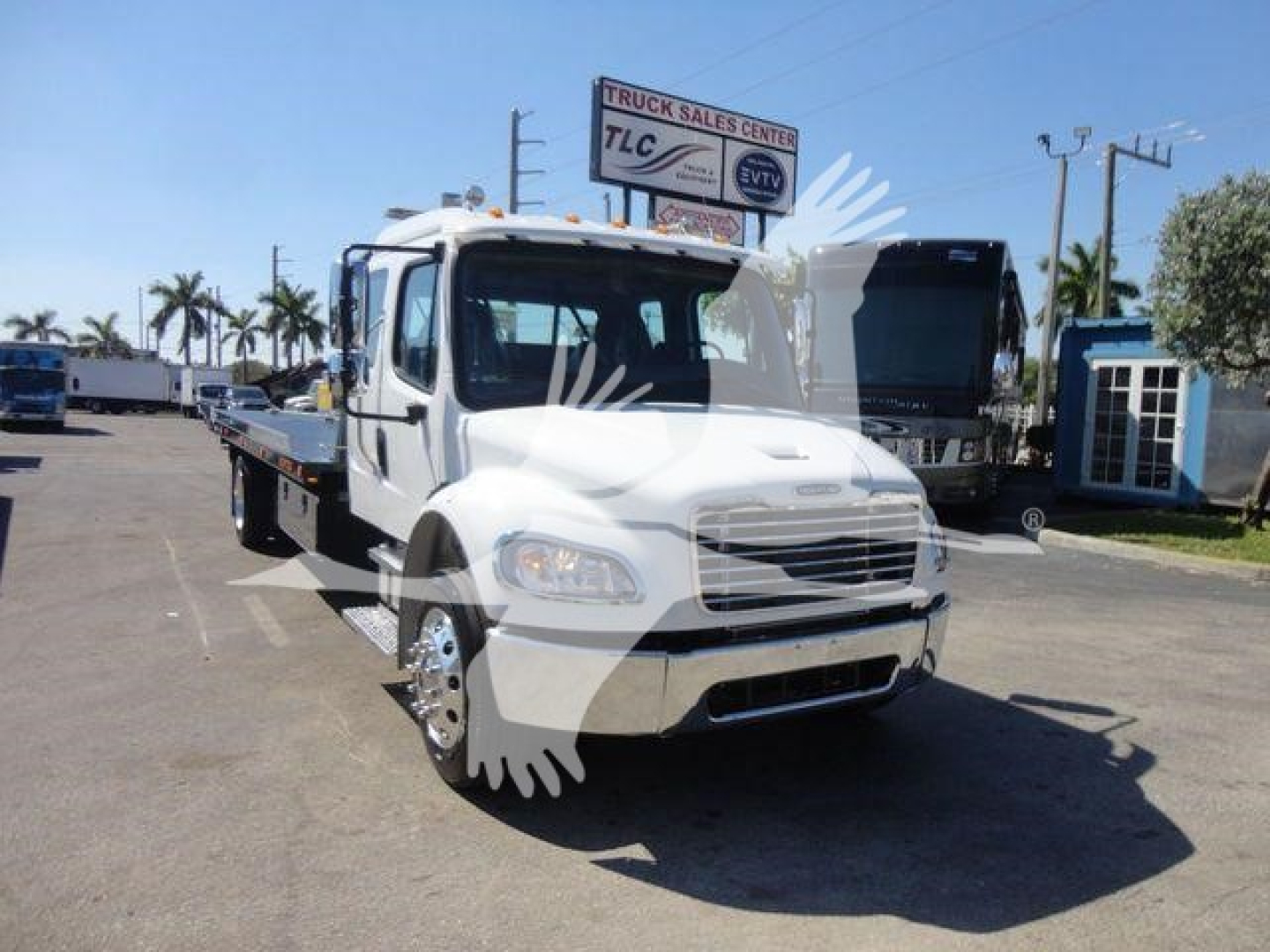 Flatbed tow truck cab in white with front grille and headlights visible, Freightliner-style hood, rolling stock yard in background.