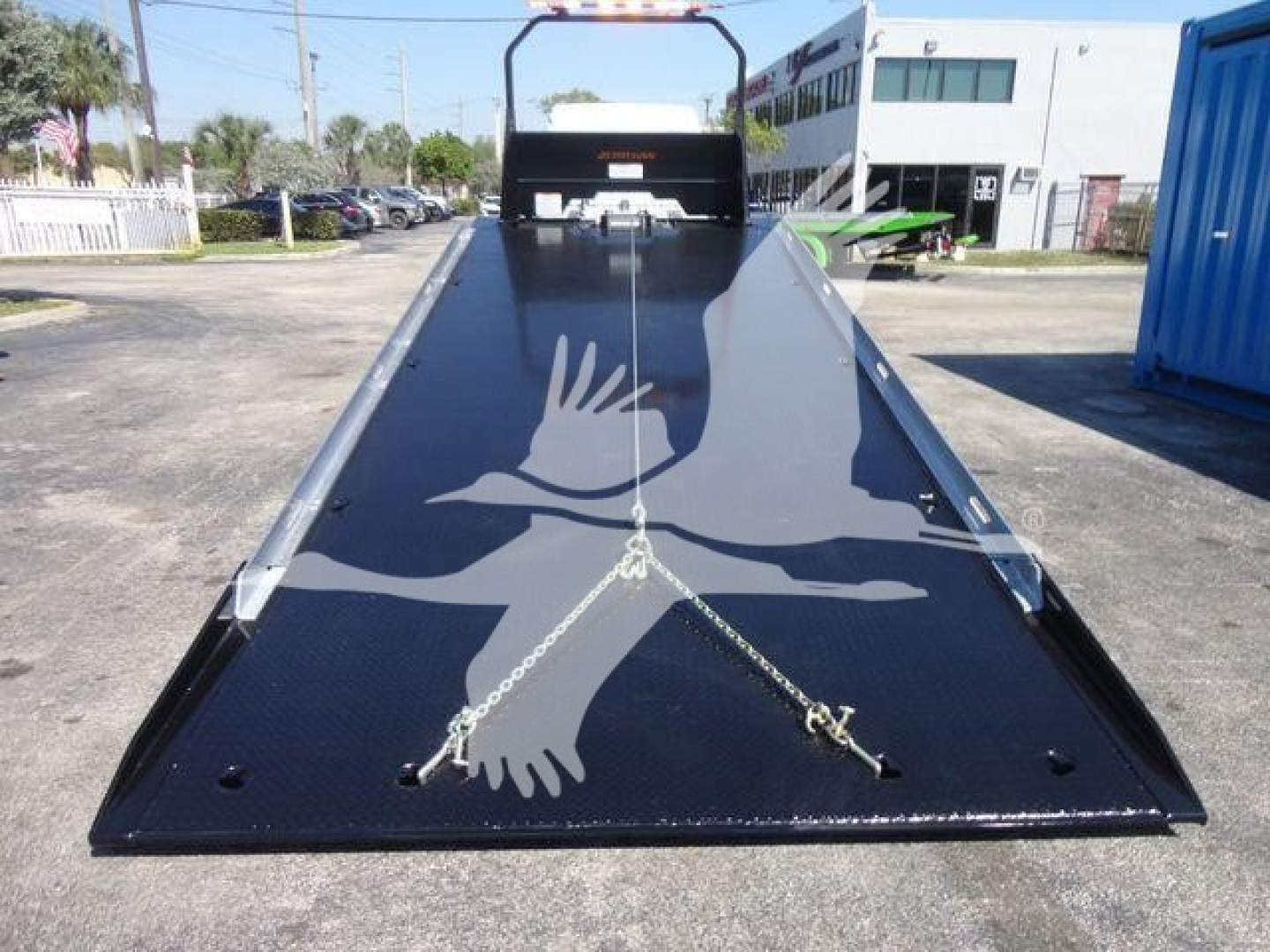 Flatbed tow truck carrier bed with black textured surface and mooring chains visible, Freightliner M2 106 equipment in lot.