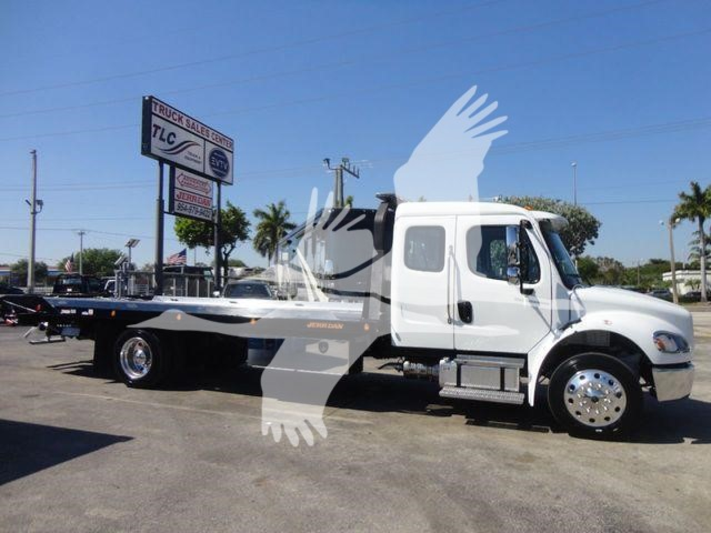 Freightliner M2 106 rollback truck in white with flatbed, side view showing cab and dual rear wheels, stock photo with dealership sign in background.