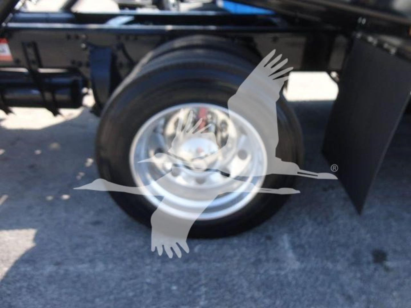 Wheel and part of Freightliner M2 106 rollback chassis showing polished steel rim and tire with shadowed undercarriage components.