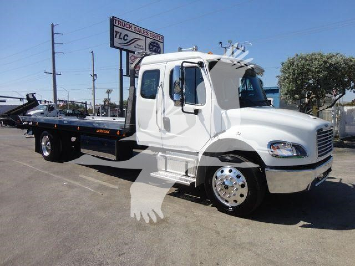 Flatbed rollback tow truck with white cab and black flatbed, Freightliner badge not visible, no additional readable text on body; background shows dealership lot and sign.