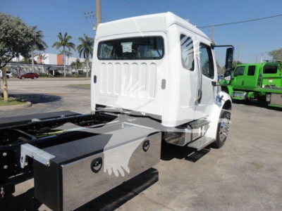 Truck cab and chassis of a Freightliner M2 106 rollback unit in a dealership lot, white cab with black chassis, side view showing sleeper cab window and rear cargo area.