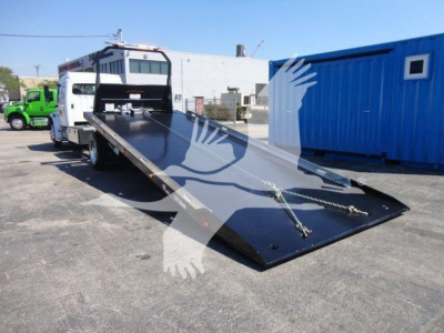 Flatbed tow truck with extended ramp on a Freightliner M2 106 chassis, black steel surface, loading ramp lowered, surrounding industrial lot
