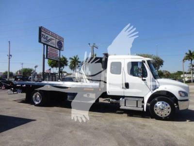 Freightliner M2 106 rollback truck in white with flatbed, side view showing cab and dual rear wheels, stock photo with dealership sign in background.