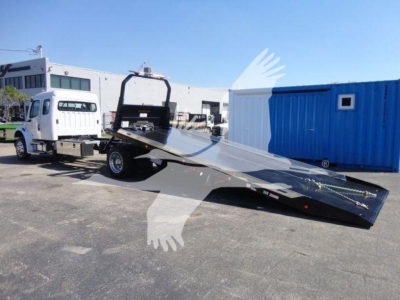 Truck with a flatbed tilt bed in a dealership lot. Visible text includes no legible labels on the vehicle itself due to distance; background shows a white cab, blue portable building.