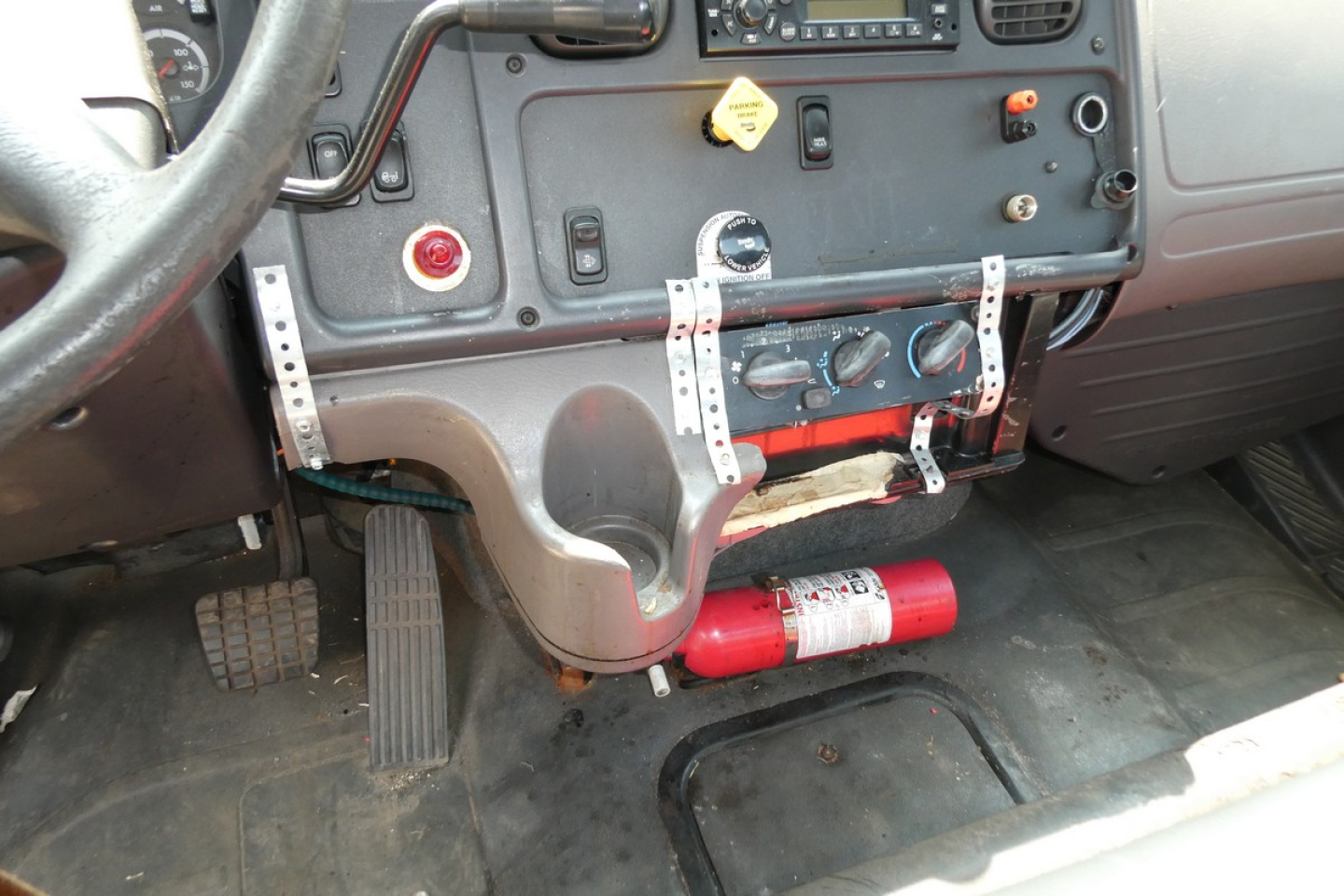 Cab area of a Freightliner M2-106 with dashboard controls, pedals, fire extinguisher, and support brackets; VIN label and equipment visible.