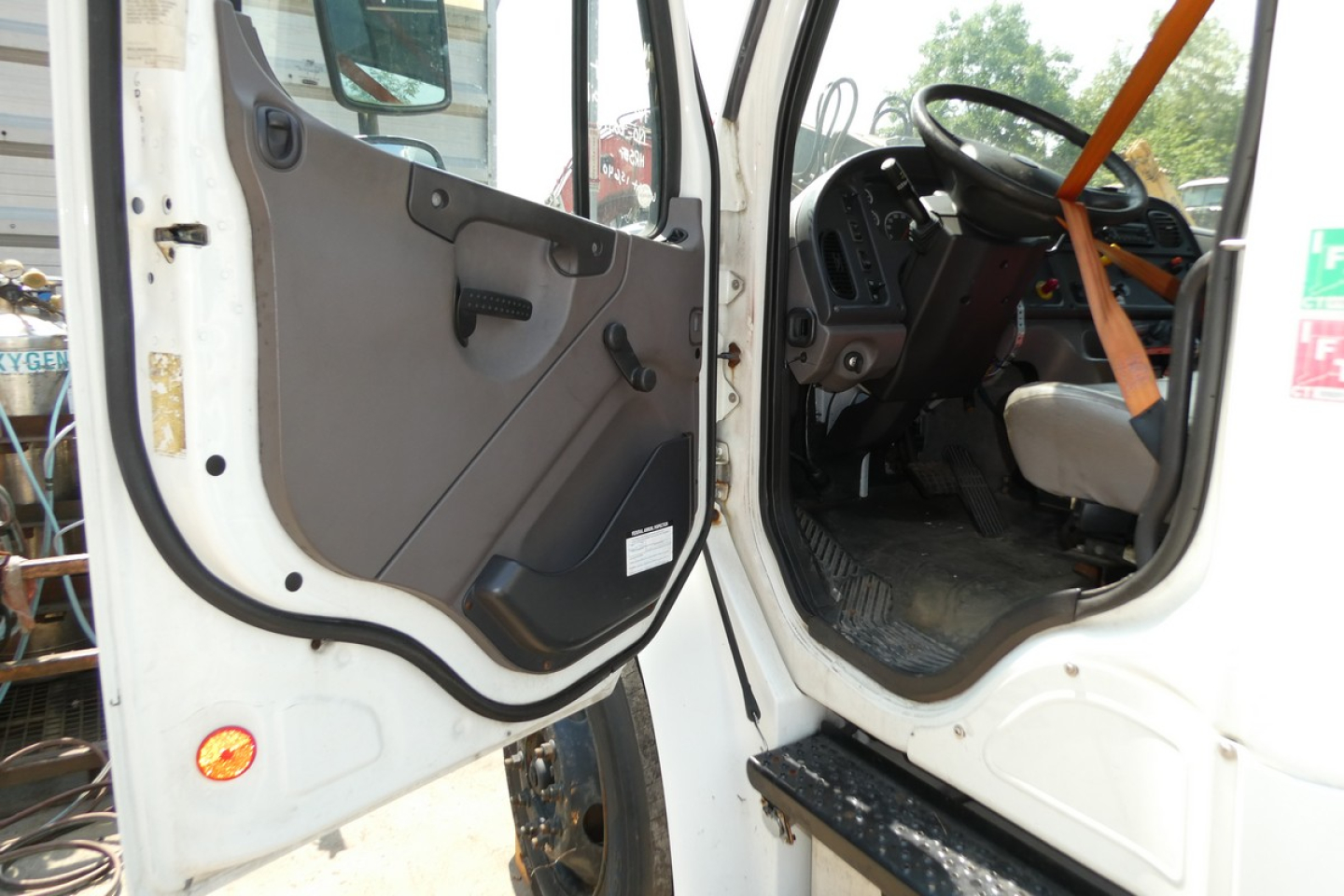 Truck cab interior and open door of a Freightliner M2-106 with steering wheel and dashboard visible.