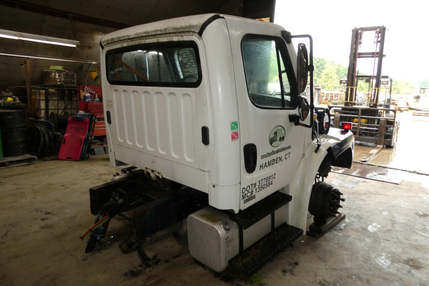 Freightliner M2-106 cab rear and left side in a shop, white body with black chassis, visible cab rear window, side mirror, and stickers.