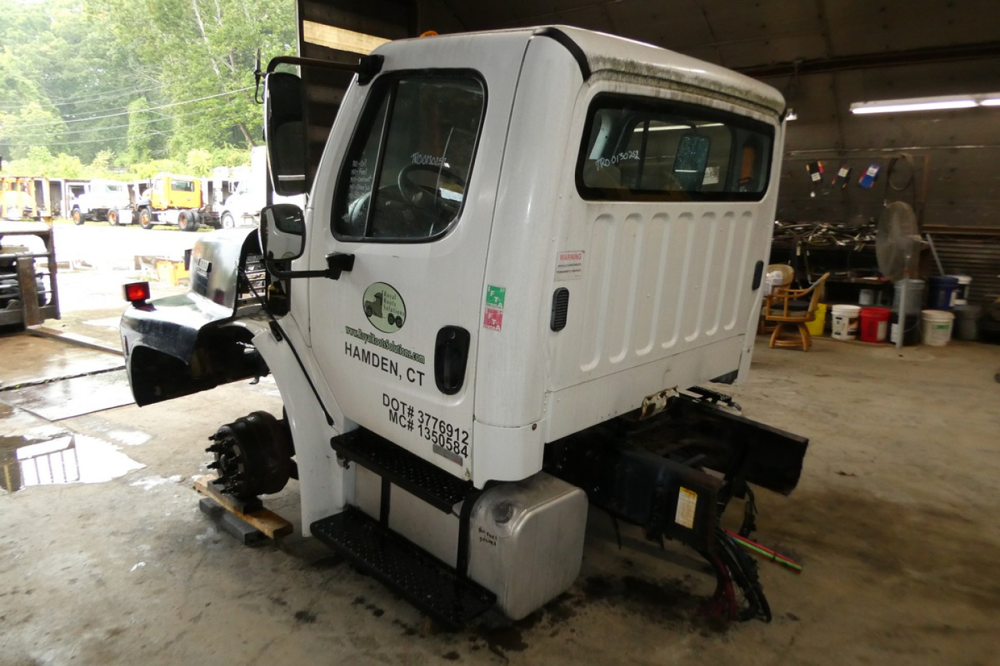 Truck cabin with missing rear section showing cab and frame; Freightliner M2-106 cab in a workshop; VIN tag and stock number on door; white exterior; no trailer attached.