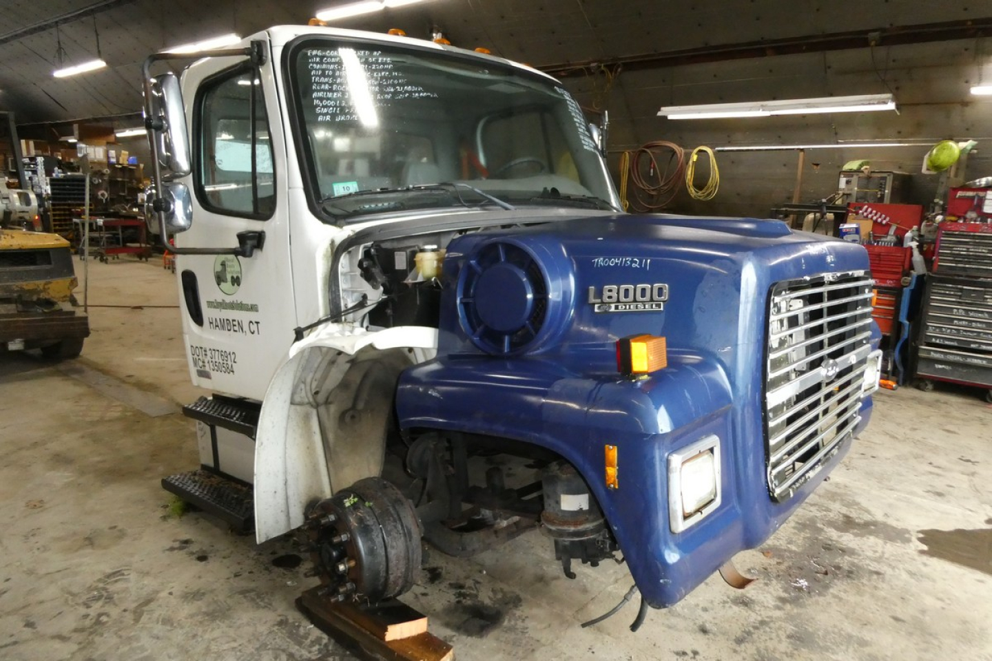 Freightliner M2-106 cab and front end with blue L8000 hood; missing front wheel, exposed brake assembly; service area in background.