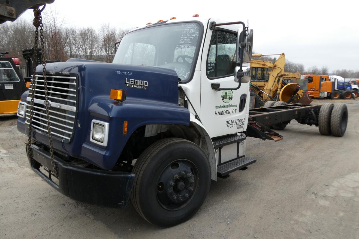 Freightliner M2-106 medium duty truck in a yard with a blue cab and white sleeper, logging chain hoist on front bumper, flatbed chassis, various orange equipment in background.