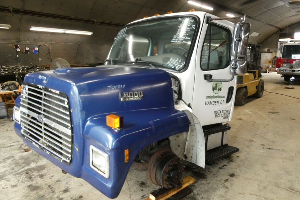 Freightliner M2-106 truck cab with blue front and white sleeper, front grille and wheel removed, inside workshop.
