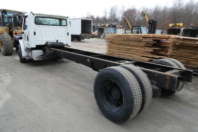 Freightliner M2-106 chassis cab showing dual rear axles and long wheelbase, white cab with flatbed/trolley, pallets in background.