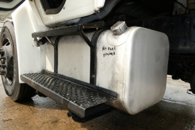 Truck side view showing a rectangular white plastic fuel tank with a black metal step and a text reading 'No fuel primed' near the tank cap; partial undercarriage of a Freightliner M2-106.