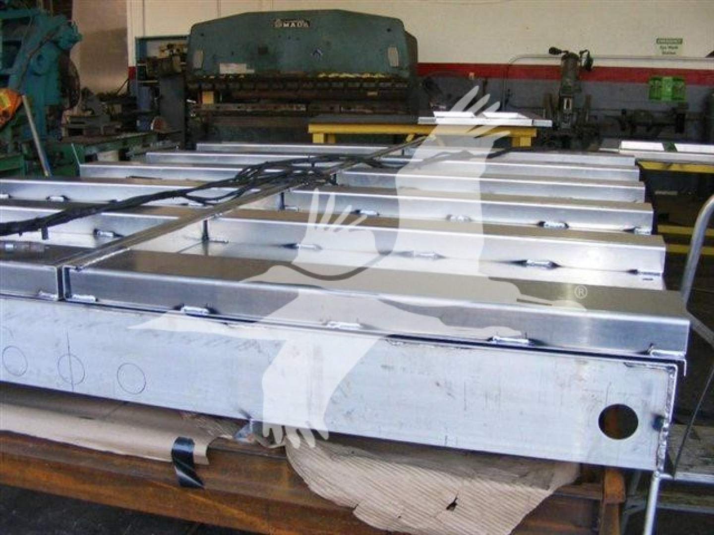 Dump truck body platform showing aluminum side rails and a large blue blue press brake in background with stock number 12TD48PS in warehouse.