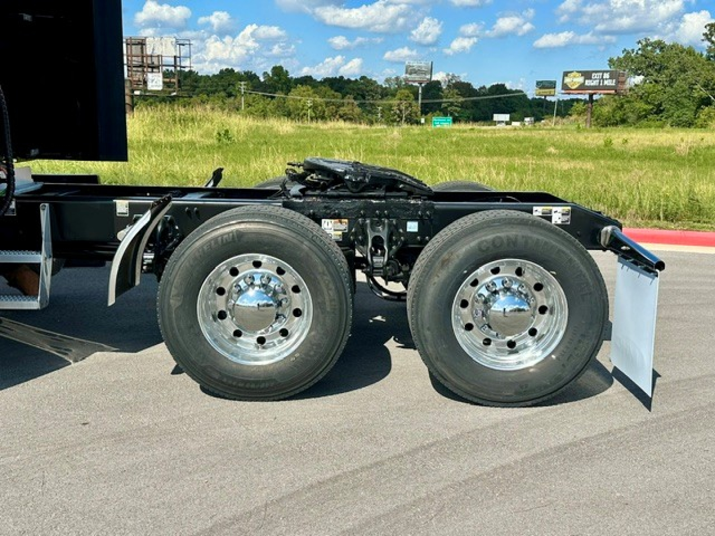 Dual rear wheel assembly of a Peterbilt semi-truck chassis with chrome hubs and tire treads visible; part of fifth wheel and frame assembly, road in background.