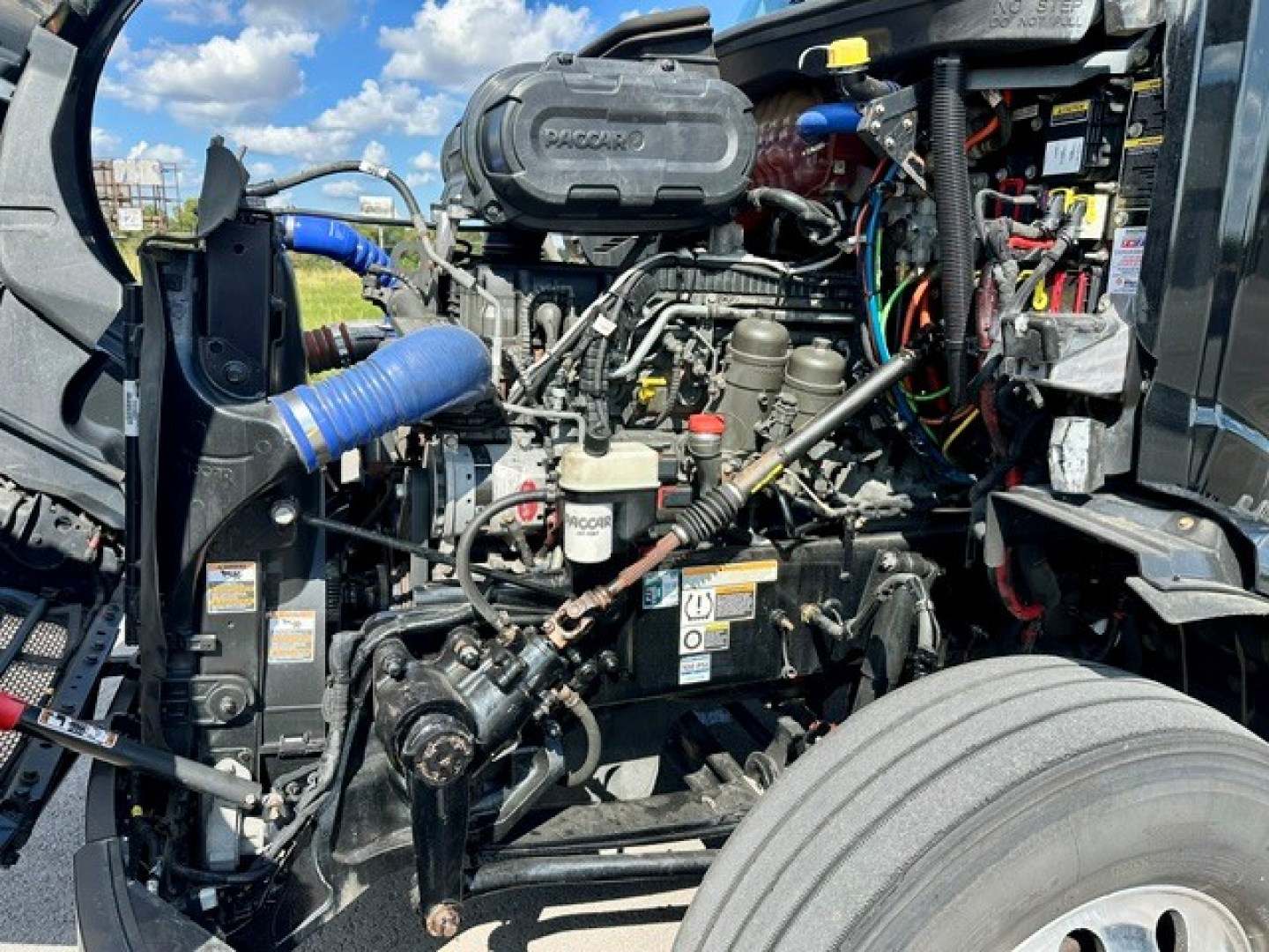 Peterbilt 579 engine bay showing Cummins or PACCAR engine components with hoses, wiring, and air intake; visible radiator, belts, and front wheel.
