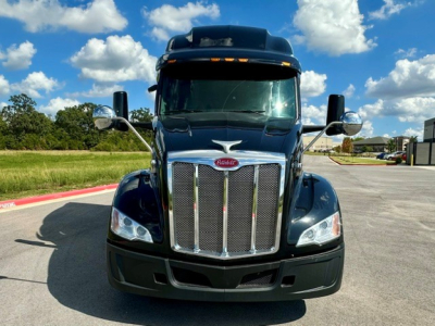 Peterbilt 579 truck facing camera, chrome grille, black body, tall sleeper cab, clear blue sky with clouds.