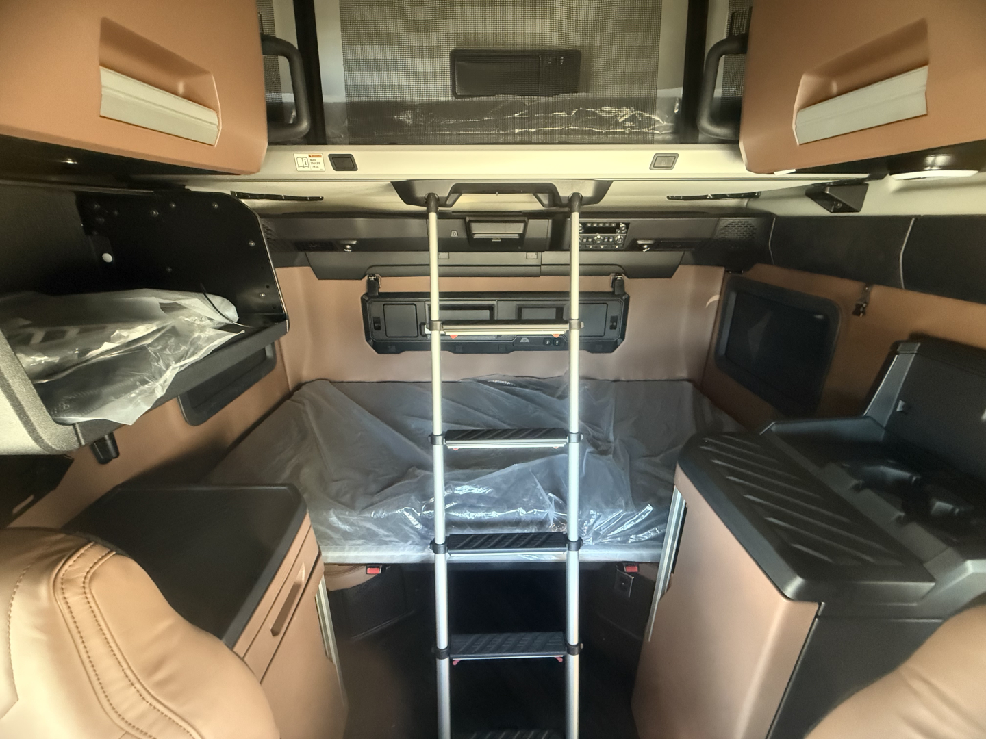 Cab interior of a new Mack PR64T76H sleeper with ladder and bunk area, beige leather accents, upper bunk, and storage compartments visible.
