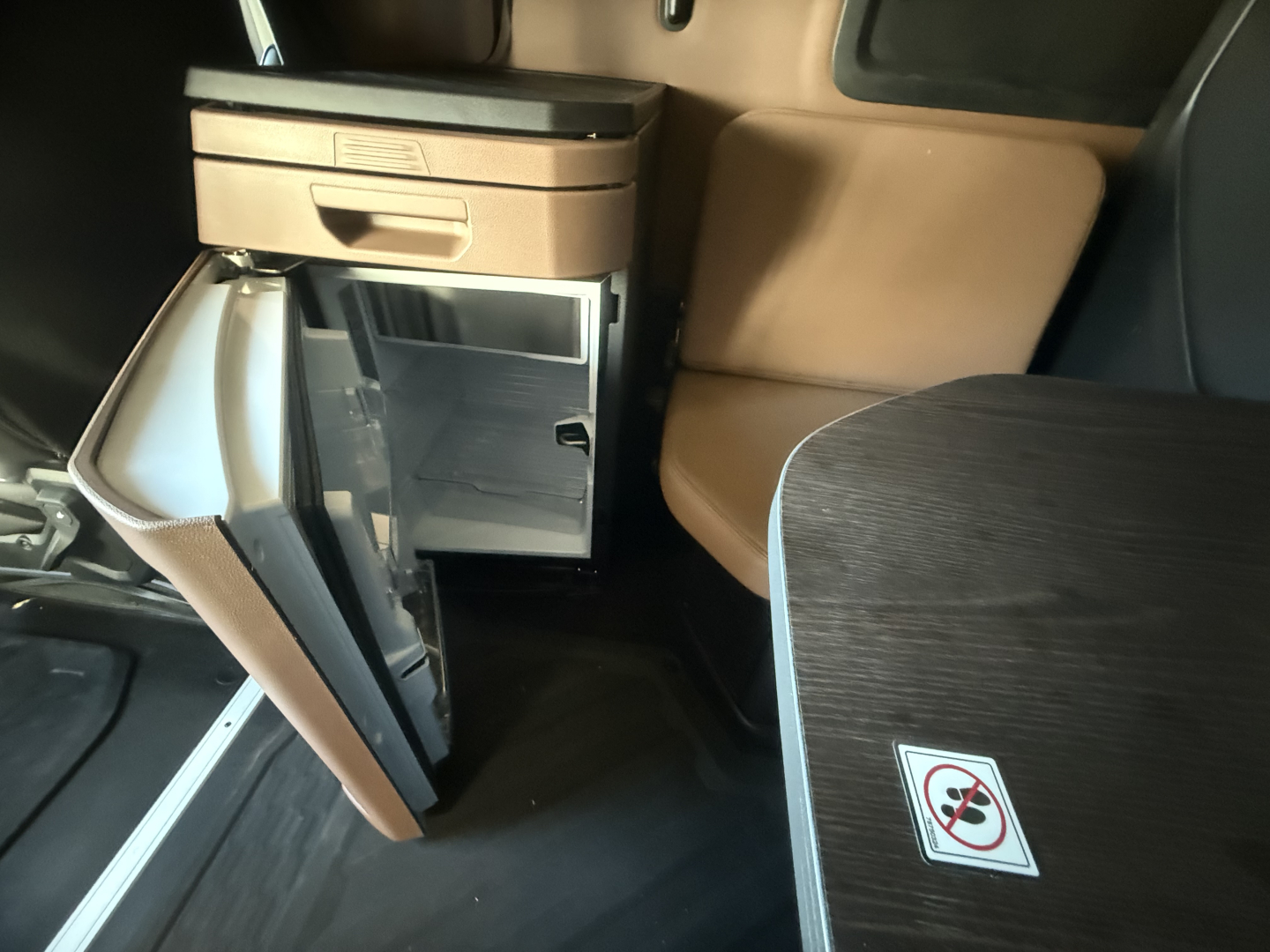 Interior of a Mack PR64T76H sleeper unit showing a small refrigerator cabinet door open and storage drawers, with a woodgrain table surface and leather seats.