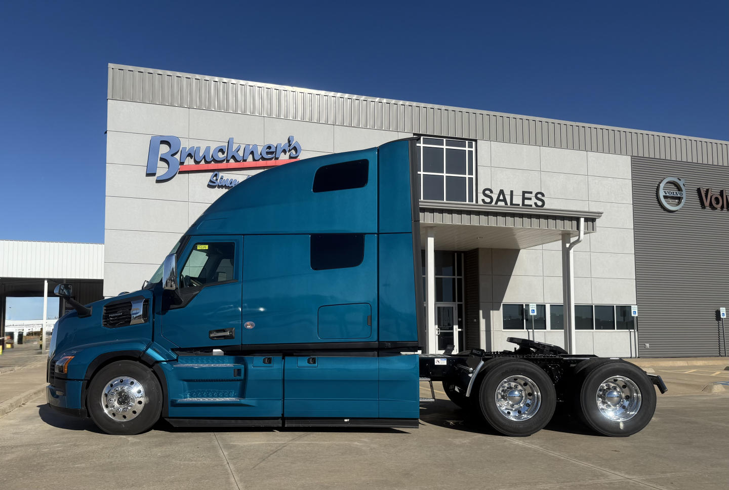 Blue Mack PR64T76H tractor in front of dealership with 'SALES' sign and a building showing Bruckner's branding.