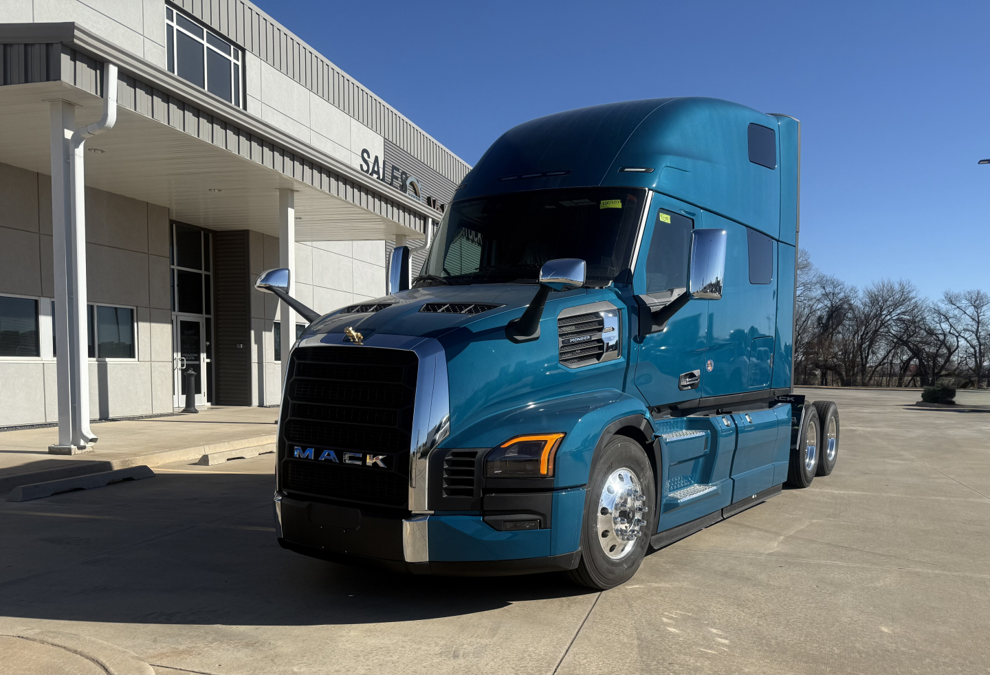 Blue Mack truck PR64T76H in-service to show front grille with MACK lettering and headlights; sleeper cab with tinted windows; dealership building in background.