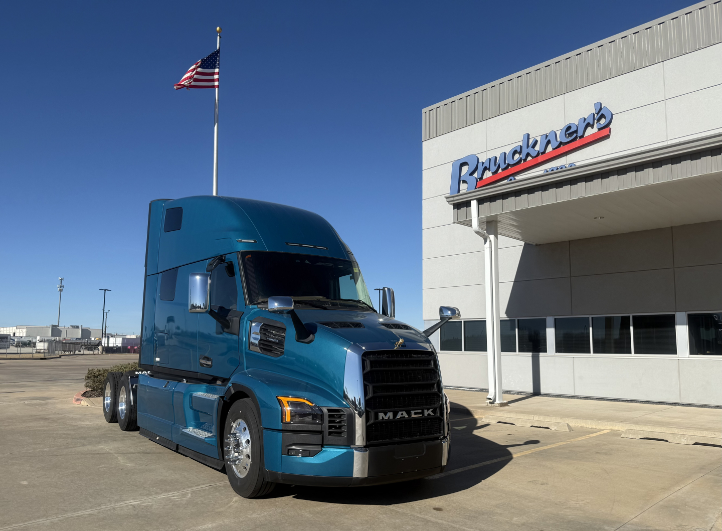 Mack PR64T76H conventional sleeper truck in teal blue parked in front of a dealership with Mack badge on grille.