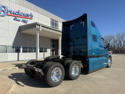 Blue Mack PR64T76H highway tractor in a parking lot, side and rear view showing cab with sleeper, dual rear axles, chrome wheels, and fifth wheel area.