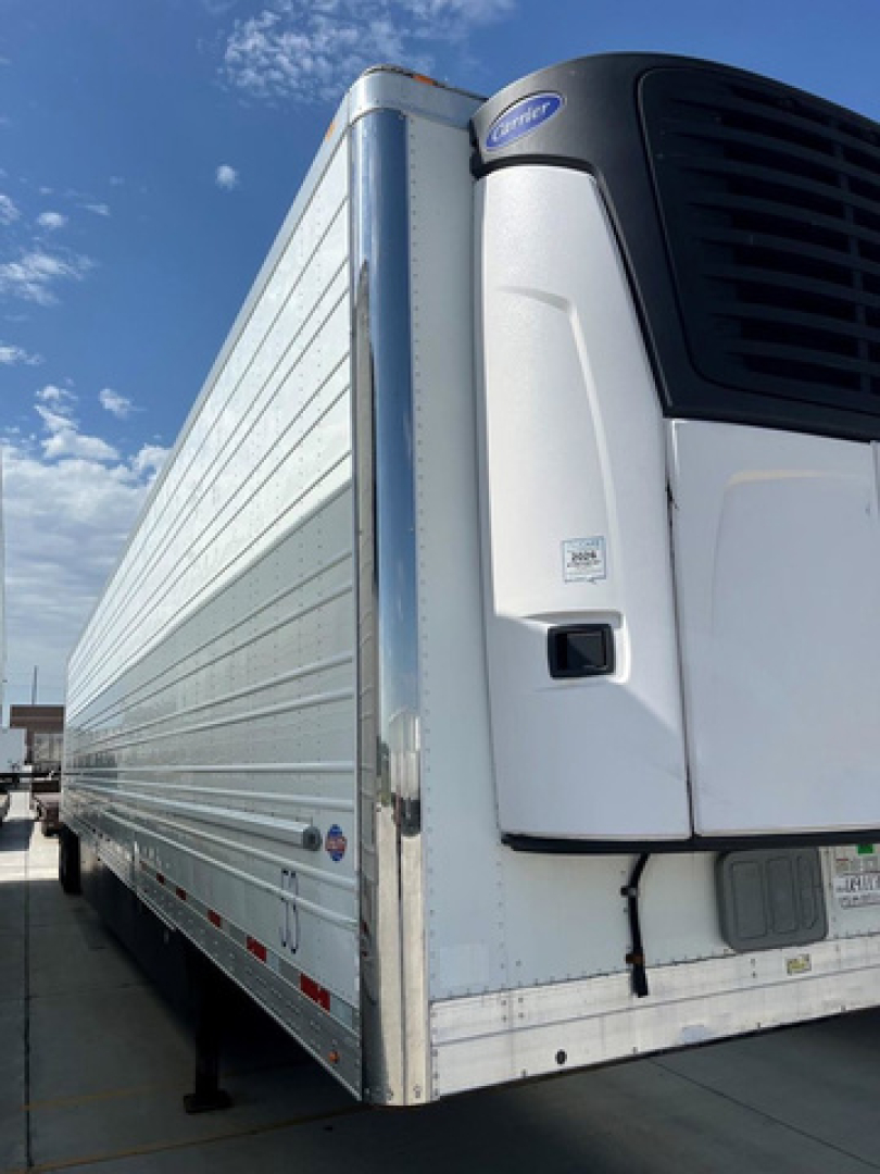 Trailer front corner with refrigerated unit on a white trailer. Visible text includes Carrier reefer unit name on the front, small labels on the unit doors, and some reflective stripes along the side.