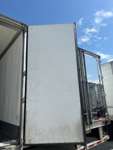 Trailer side door of a refrigerated Utility trailer, plain white surface with open rear doors and chrome hinges, blue sky in background.