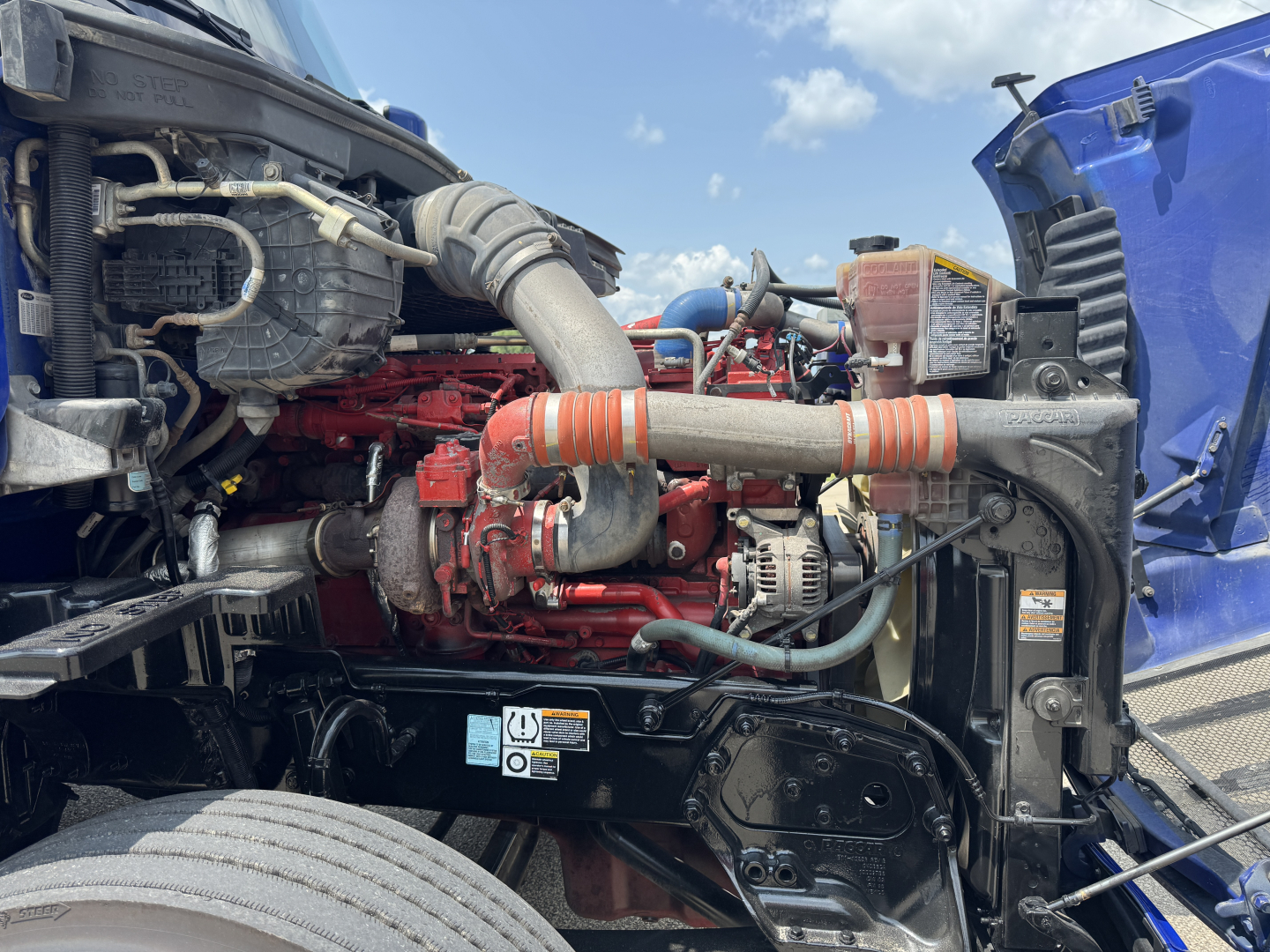 Peterbilt 579 truck engine bay with red engine, orange intake hose, black frame, and blue cab visible; various hoses and components; VIN sticker not legible.