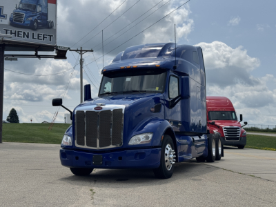 Peterbilt 579 semi-truck in blue, front view, with chrome grille and sleeper cab. A red truck is in the background. Billboard sign partially visible on left.