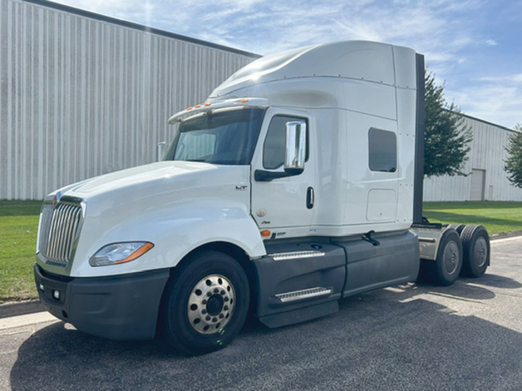 Truck cab in-view showing International LT625 with sleeper cab and dual rear axles, white exterior, front grille, and side steps.