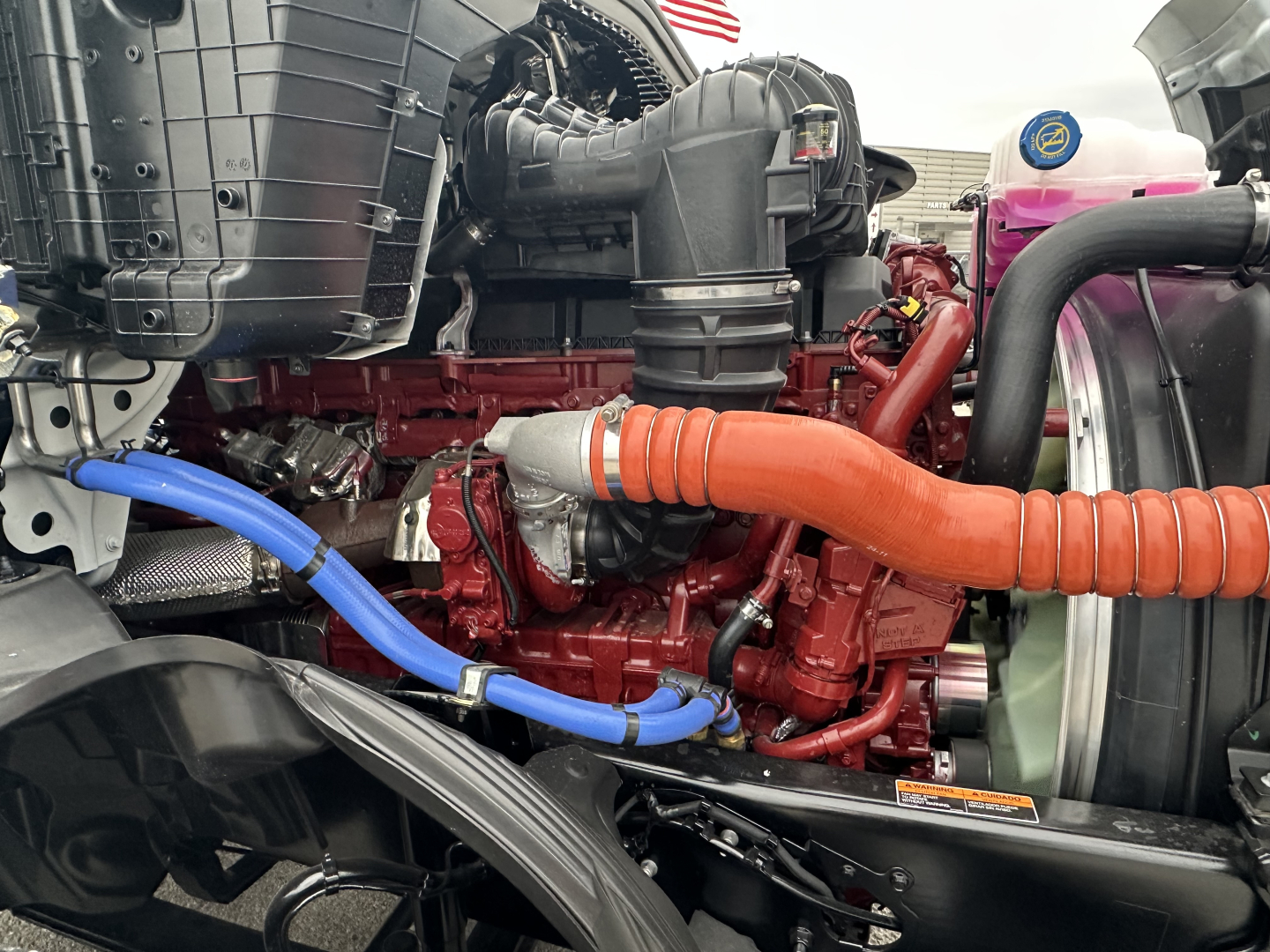 Mack PR64T engine bay showing red Cummins/engine component with blue and orange hoses, air intake, radiator, and surrounding black plastic components.