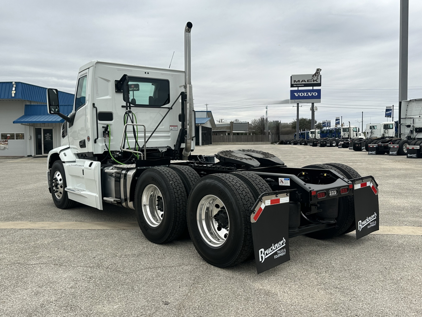 Truck chassis of a 2026 Mack PR64T daycab shown from a rear three-quarter view, with dual rear axles, fifth wheel, and black mud flaps reading Brockton or brand; white cab, dealership lot in background.