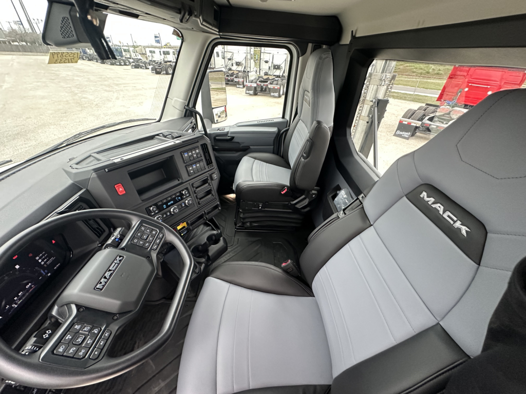 Cockpit view of a new 2026 Mack PR64T truck showing steering wheel, dashboard, and grey/black seats with Mack branding.