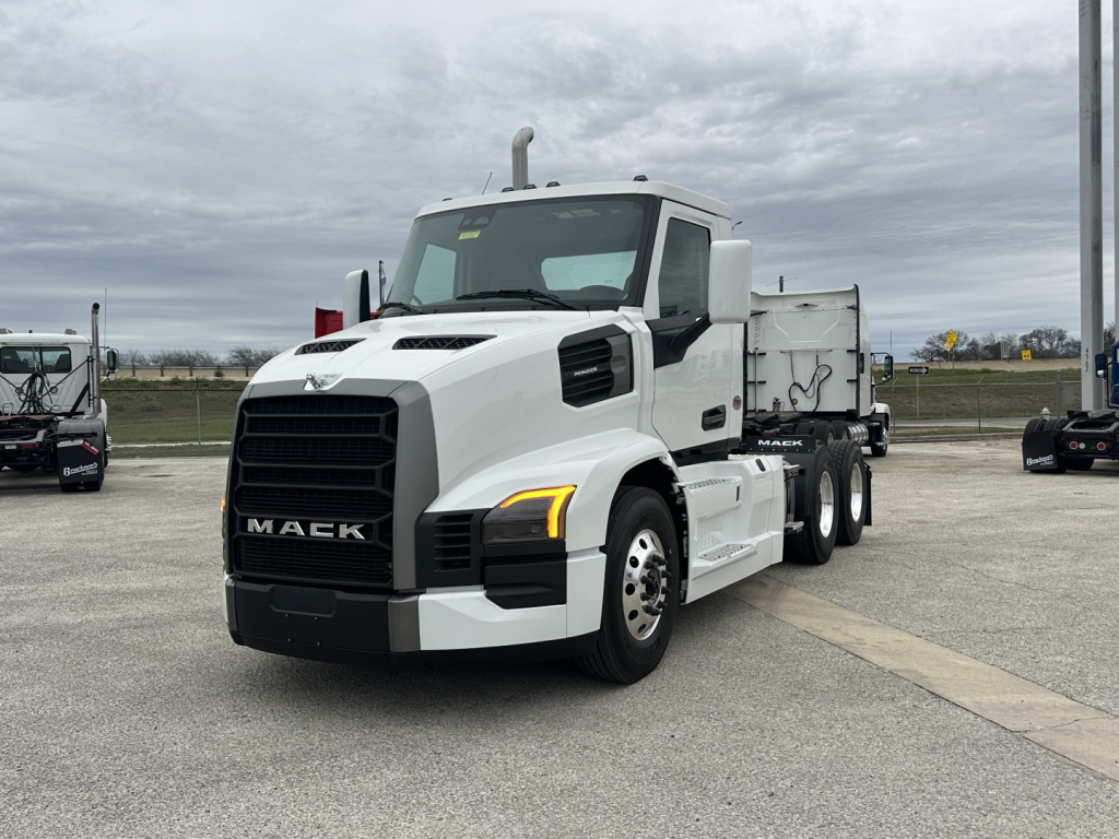 Mack PR64T truck in a dealership lot, white daycab with large black grille, yellow front turn signal, side mirrors, and a gray dump trailer behind.