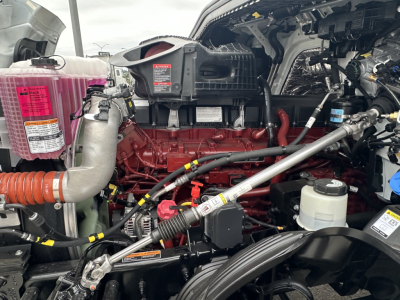 Engine bay of a 2026 Mack PR64T showing red inline engine, pink coolant reservoir, various hoses, and metal brackets.