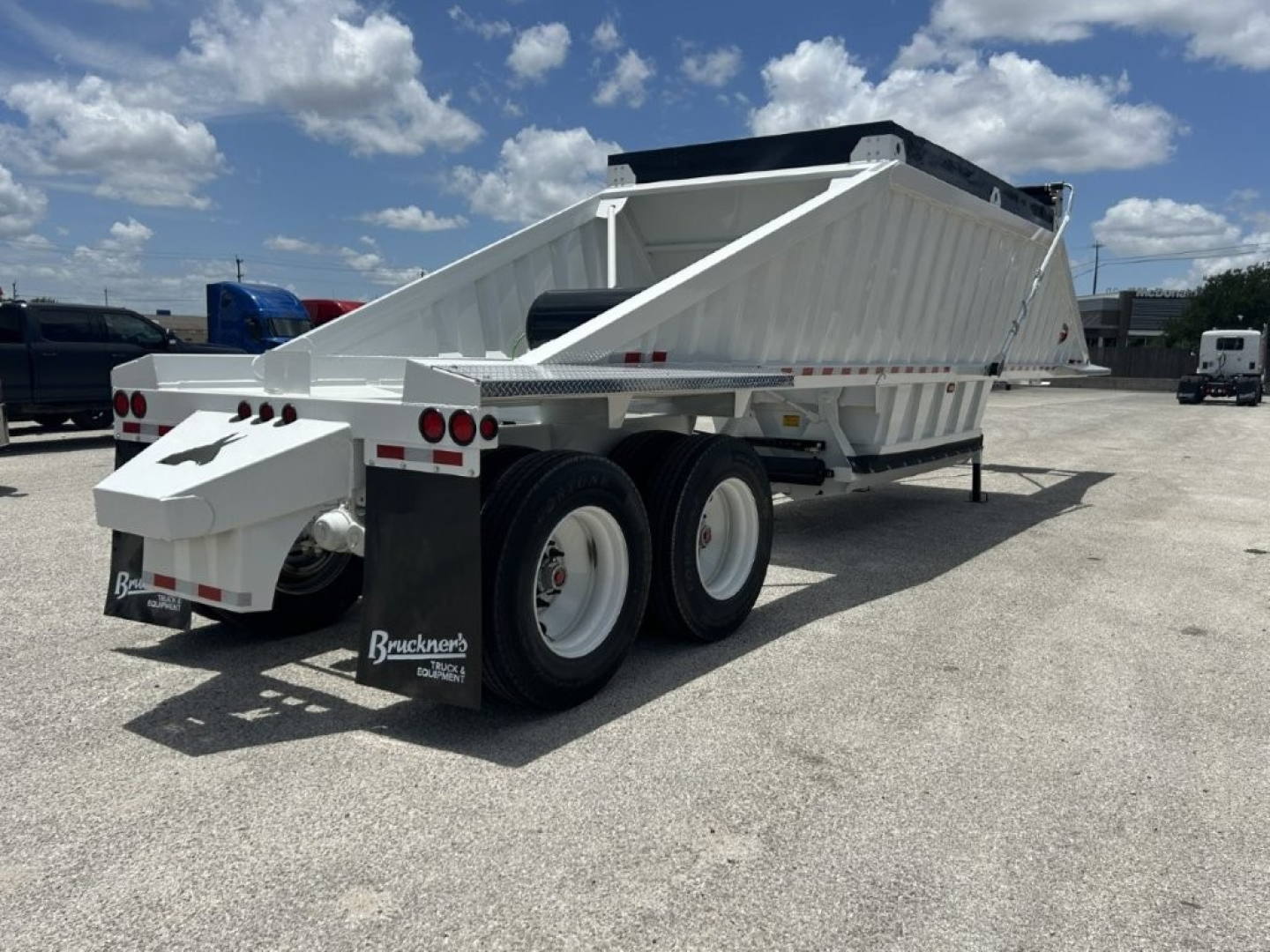 Semi-trailer dump trailer with white body and dual rear axles, viewed from side rear, showing dumping bed and wheel wells; no text visible on side; BSuckner logo on mudflaps