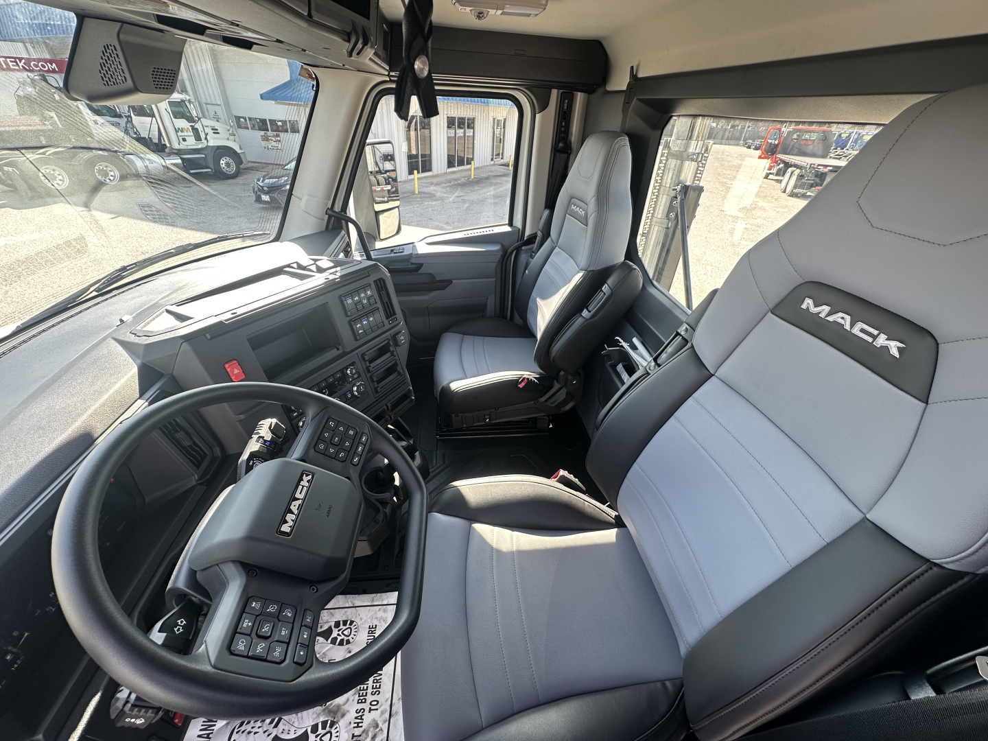 Mack PR64T truck cab interior showing steering wheel, dashboard, and gray leather seats with Mack logo.