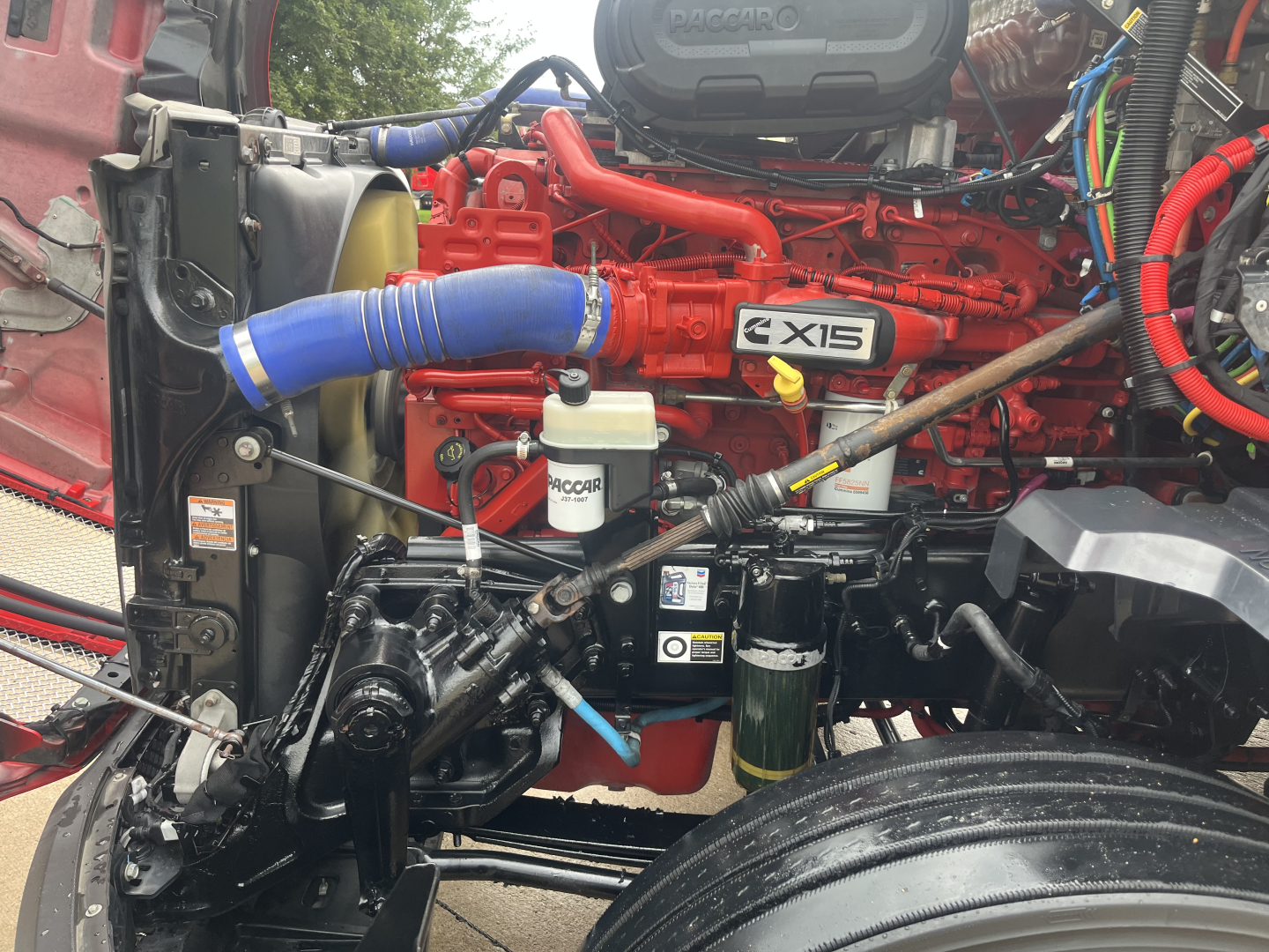 Kenworth T680 engine bay showing 6-cylinder turbo diesel with red engine block, blue intercooler hose, white AC/DEF reservoir, and various black hydraulic lines.