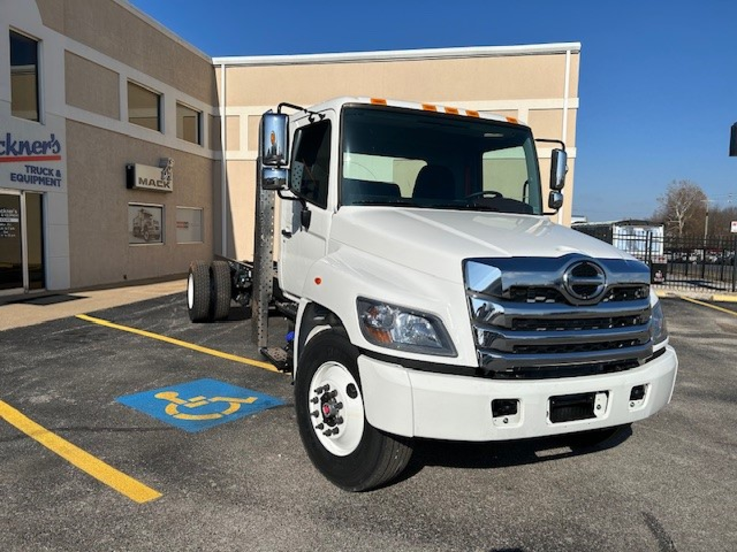 White 2025 Hino L7 cab and chassis truck in a dealership lot, front-right view showing Hino grille, bumper, and dual rear wheels.