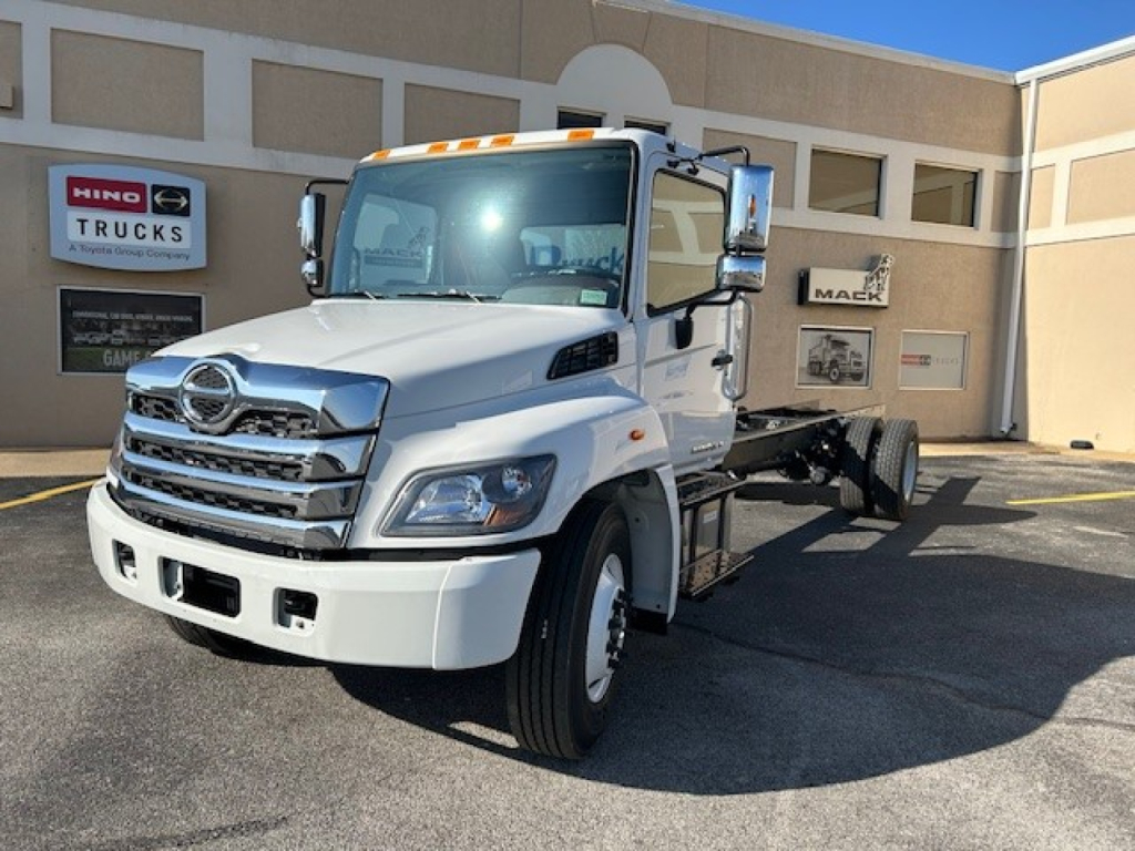Truck cab and chassis showing white 2025 Hino L7 with chrome grille and dual exhaust, front view, dealership in background.