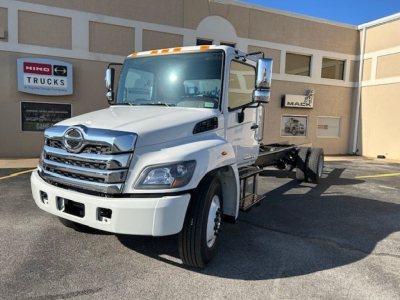 Truck cab and chassis showing white 2025 Hino L7 with chrome grille and dual exhaust, front view, dealership in background.