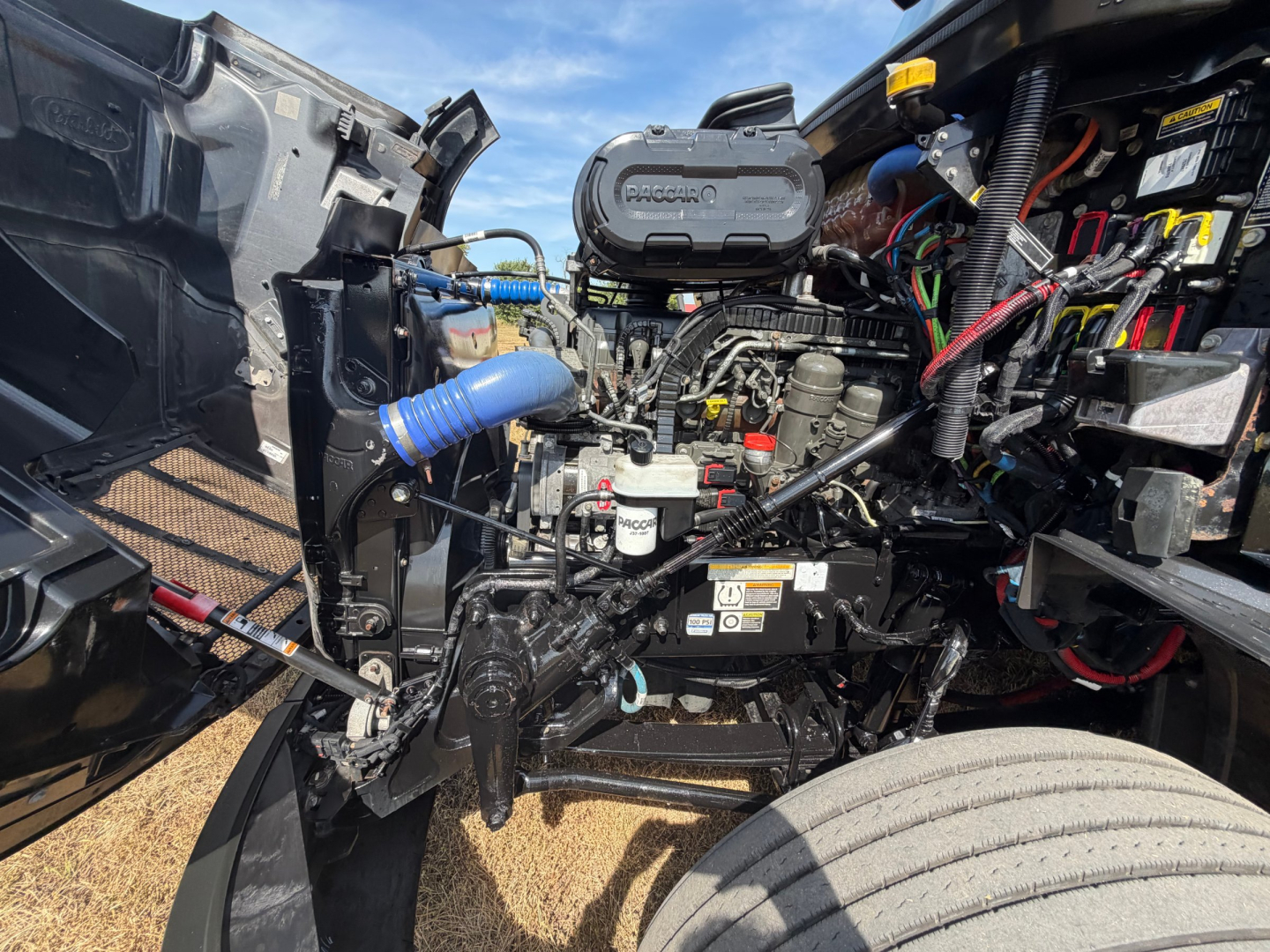 Peterbilt 579 left front engine bay with visible components including air intake tubing, battery wiring, coolant reservoir, and various labels in the engine area.