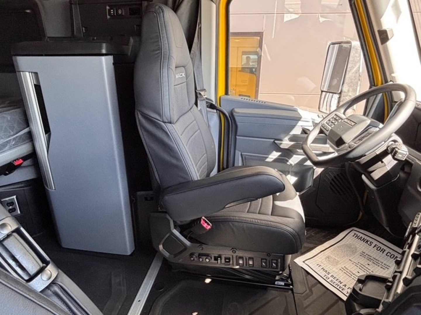 Driver seat and cab interior of a 2026 Mack PR64T76H truck showing steering wheel, dashboard, and a tall refrigerator unit in the cab.