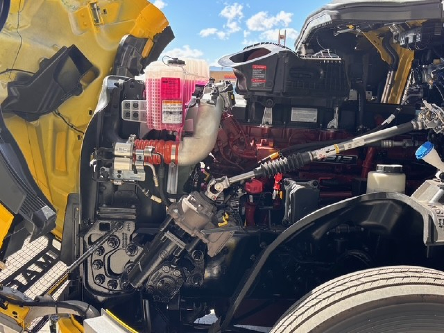 Engine bay of a 2026 Mack PR64T76H sleeper truck showing coolant reservoir, pink coolant overflow tank, hoses, and engine components.