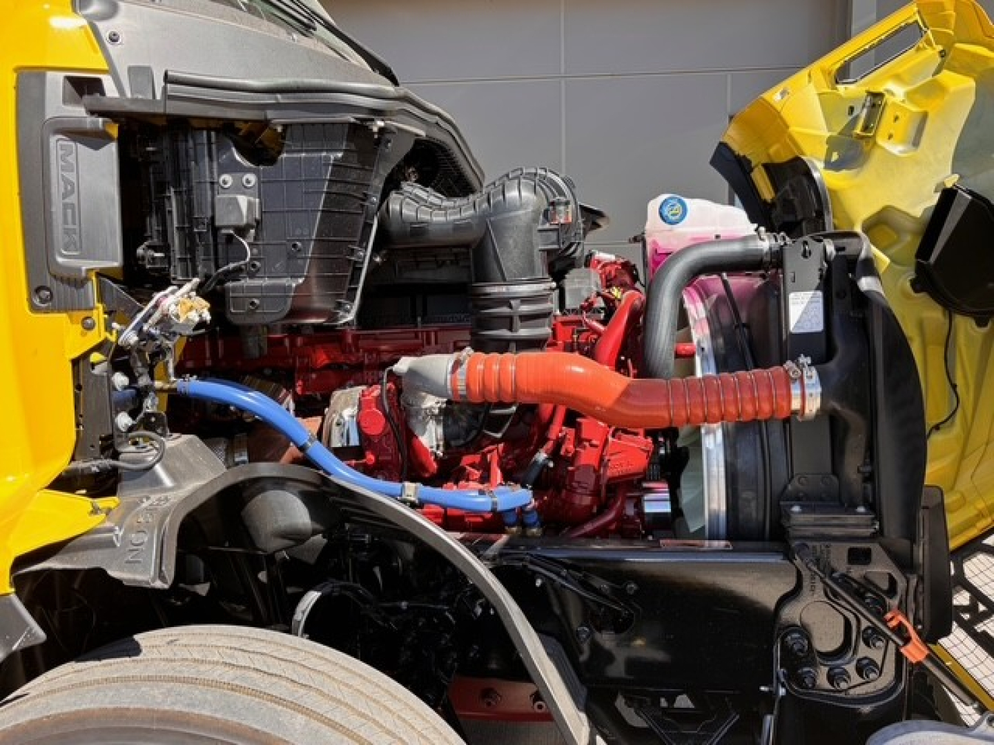 Engine bay of a Mack PR64T76H truck showing red engine block, blue hoses, orange coolant hose, black radiator, and yellow cab frame area.