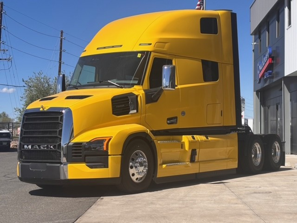 Yellow Mack PR64T76H sleeper tractor with front grille, headlights, and side mirrors, three rear axles, parked outdoors.