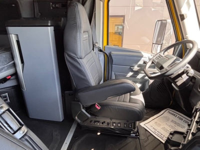 Driver seat and cab interior of a 2026 Mack PR64T76H truck showing steering wheel, dashboard, and a tall refrigerator unit in the cab.